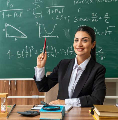 impressed-young-female-teacher-sits-table-with-school-tools-raising-pen-classroom